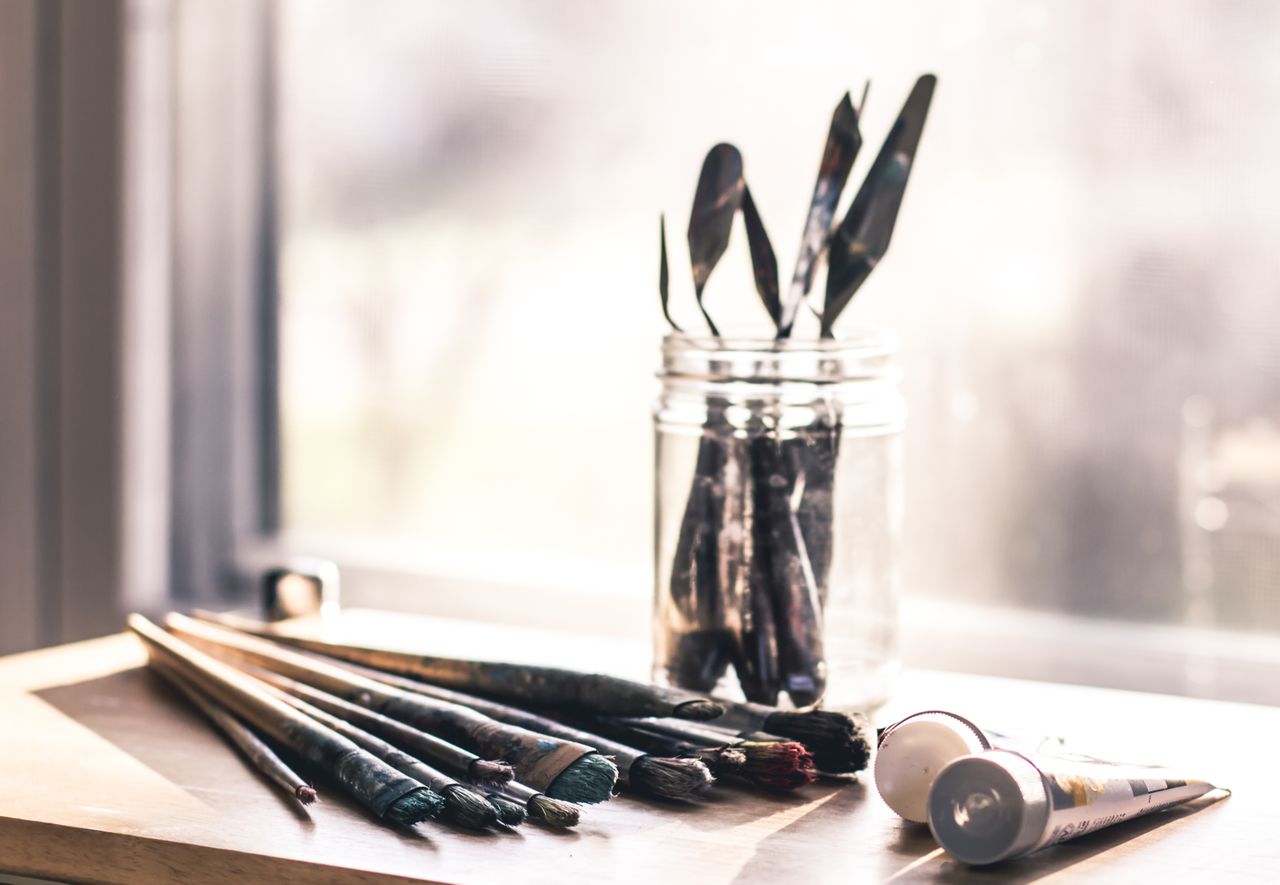 Glass jar with palette knives and paintbrushes on a sunny table, evoking creativity.